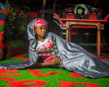 A woman in colorful traditional attire poses gracefully indoors with decorative surroundings.
