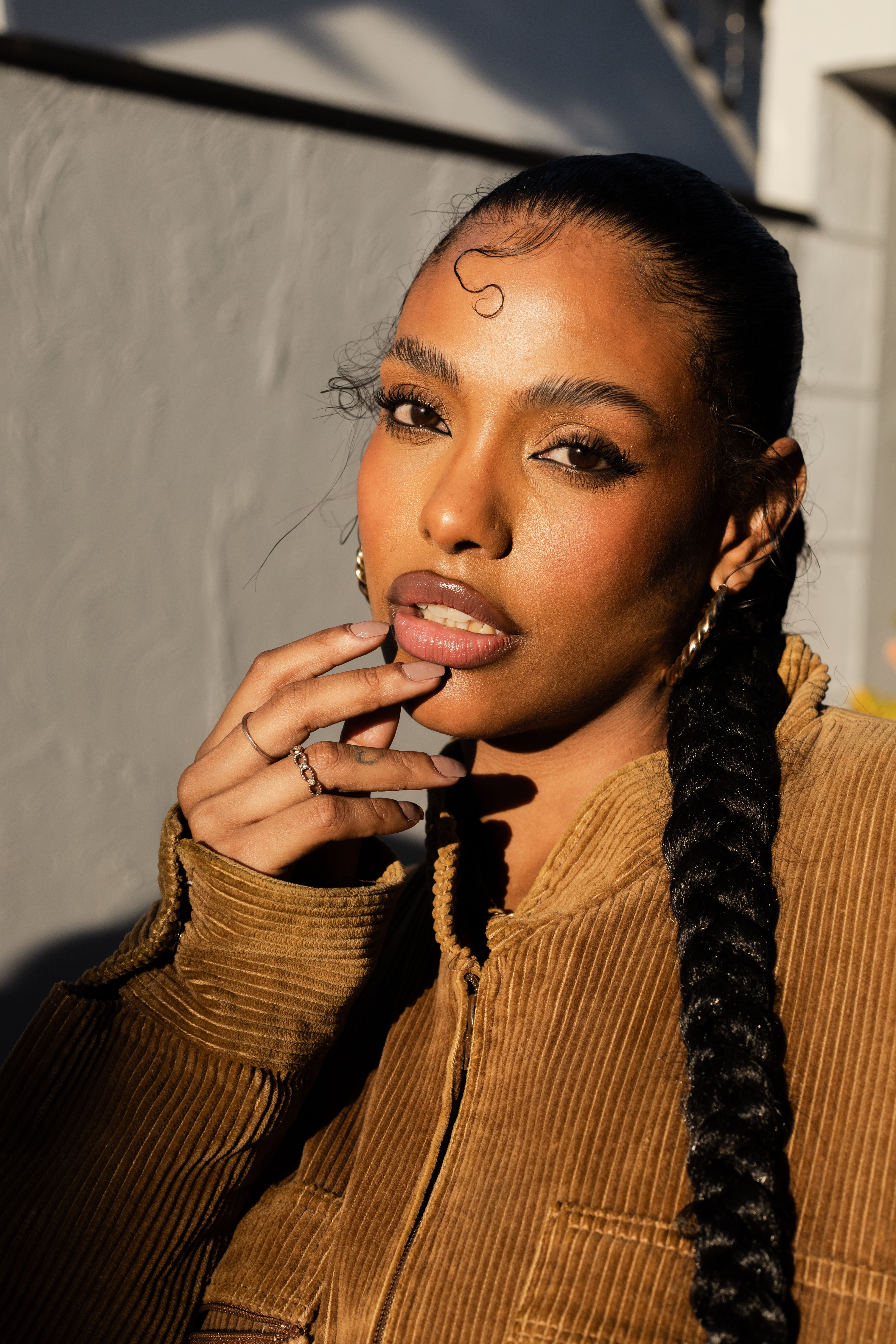 A woman with braided hair poses confidently in sunlight, wearing a brown jacket. Fashion portrait in São Paulo.