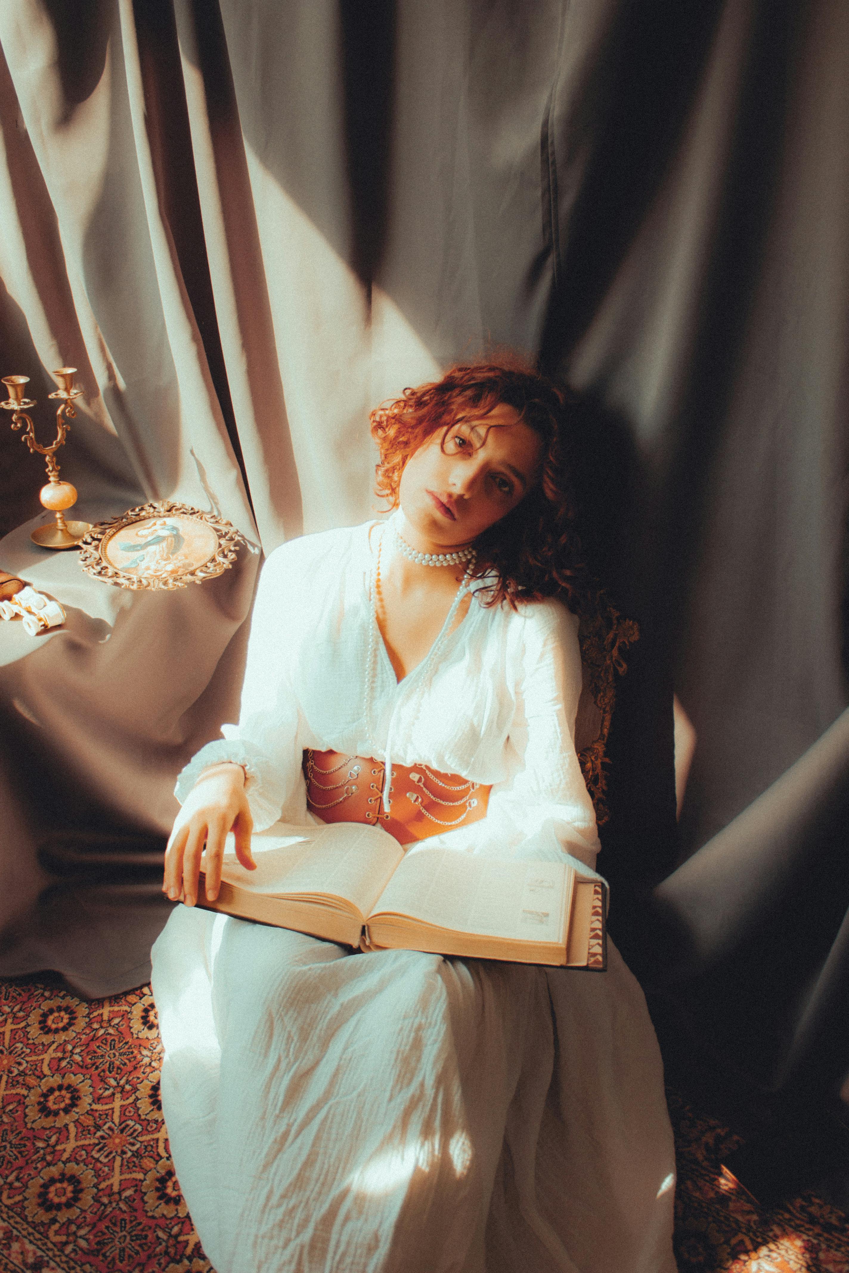A woman in a white dress reads a book in a soft, vintage setting with warm light.