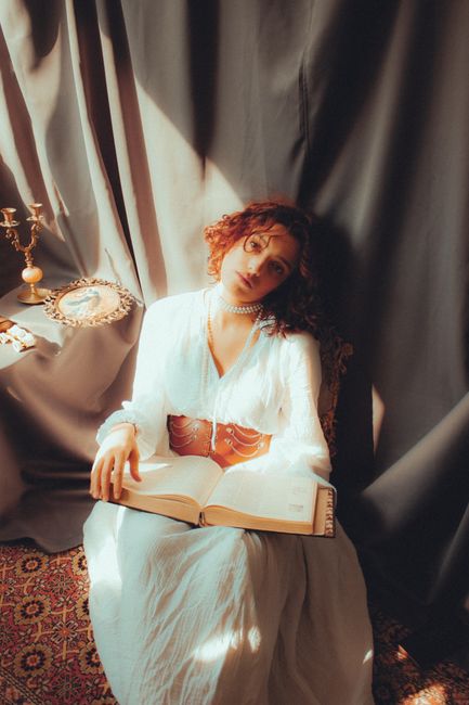 A woman in a white dress reads a book in a soft, vintage setting with warm light.