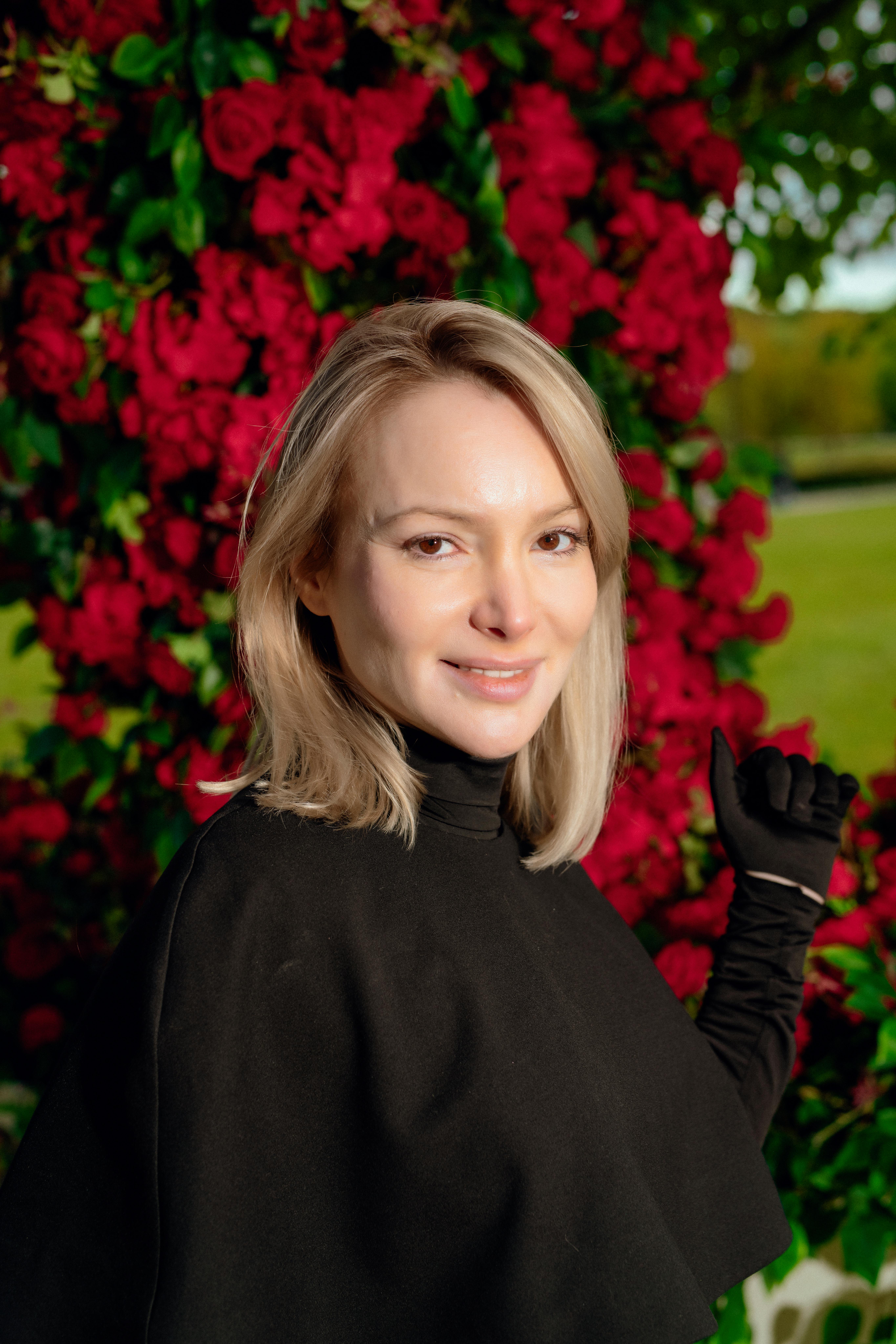 Ilmainen Hymyilevä nainen mustassa takissa poseeraa ulkona kirkkaanpunaisten kukkien keskellä. Kuvapankkikuva