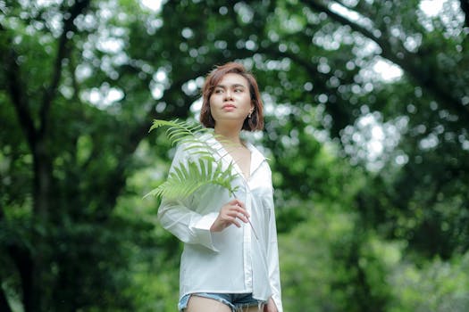 A young woman holding a fern leaf stands in a sunlit forest, surrounded by lush greenery.