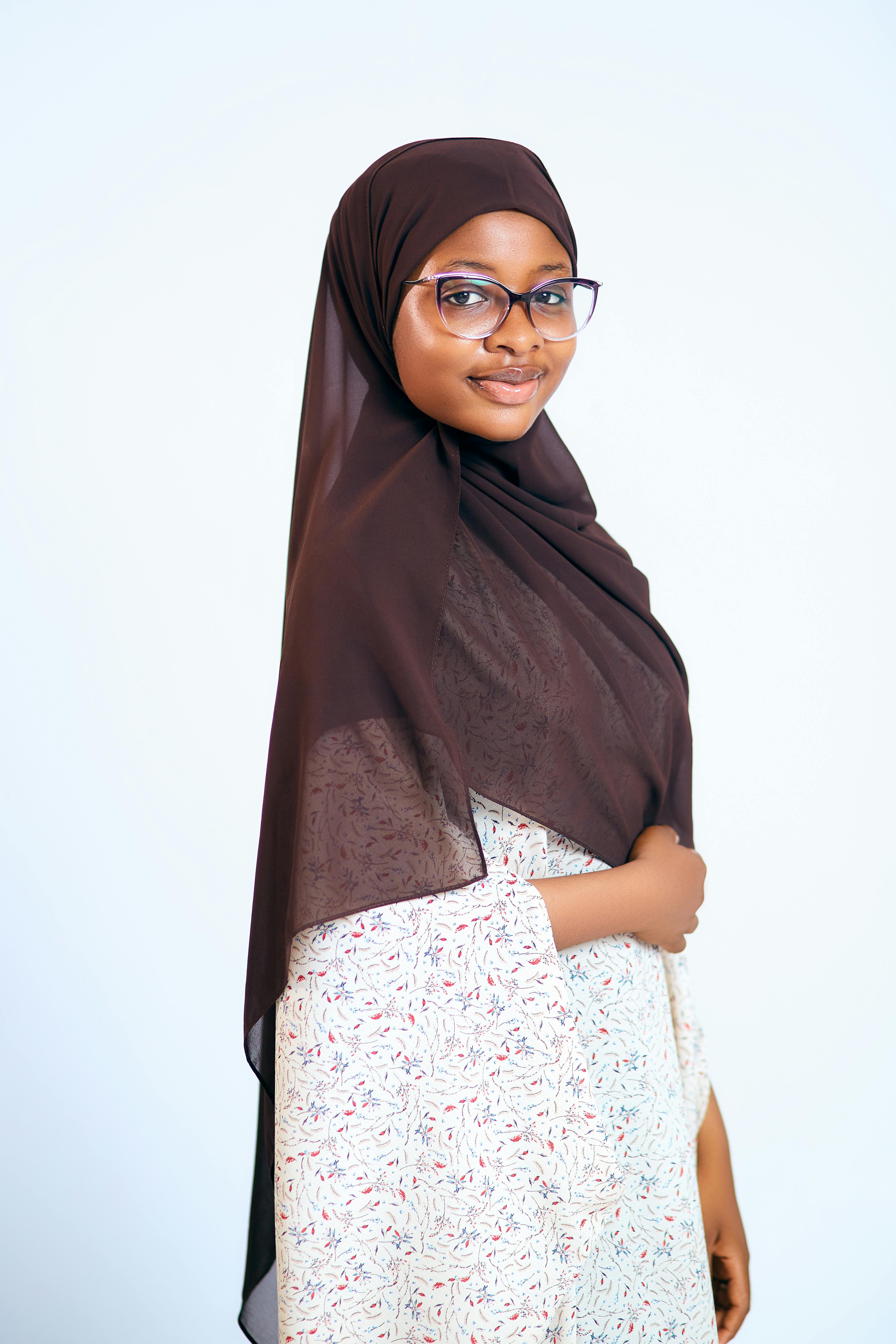 Confident young woman wearing a hijab and glasses posing against a plain background.