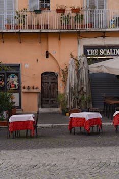 View of a quaint Italian cafe with red tablecloths and empty chairs, set outdoors on a cobblestone street.