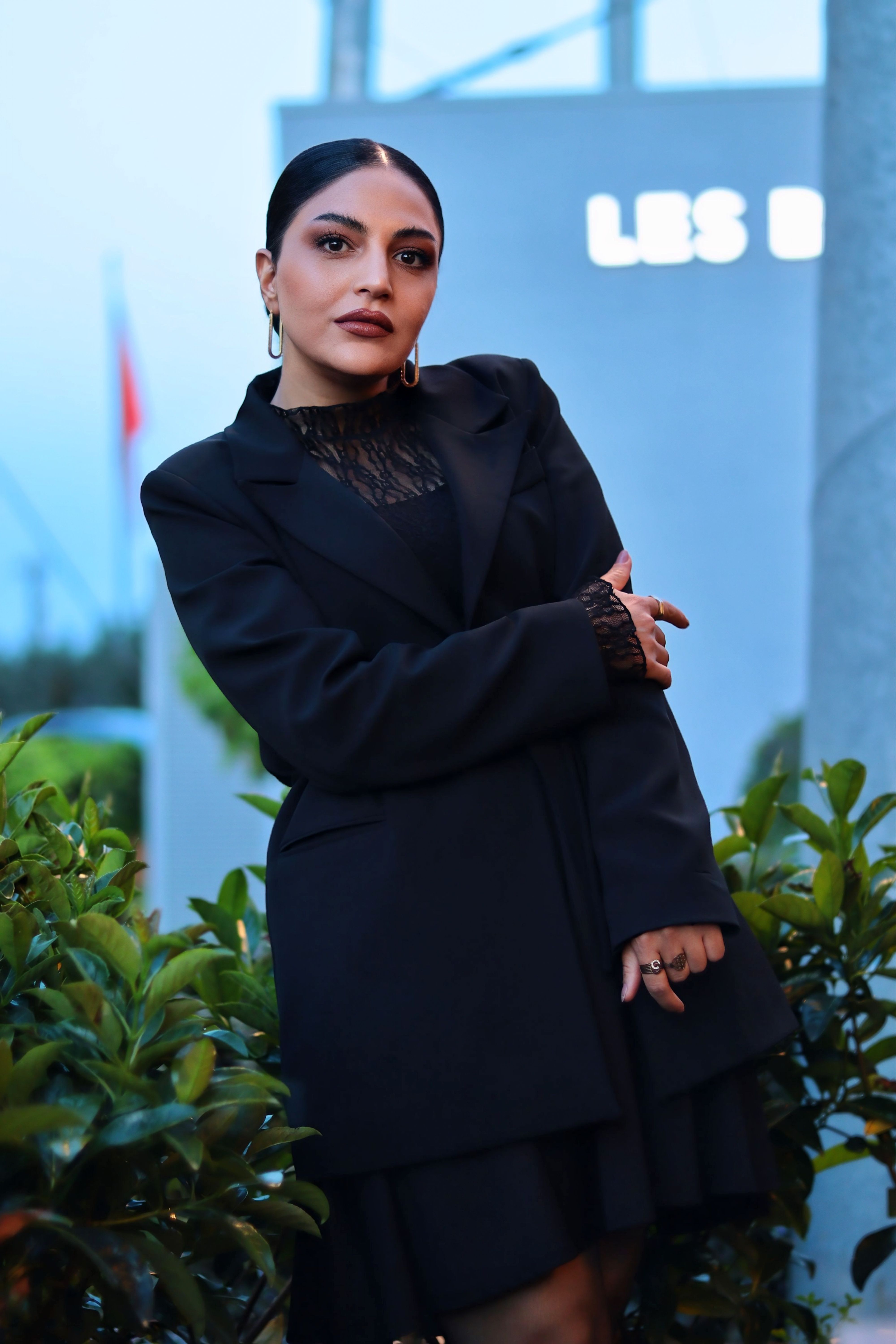 Elegant woman posing in a dark blazer amidst green foliage outdoors.