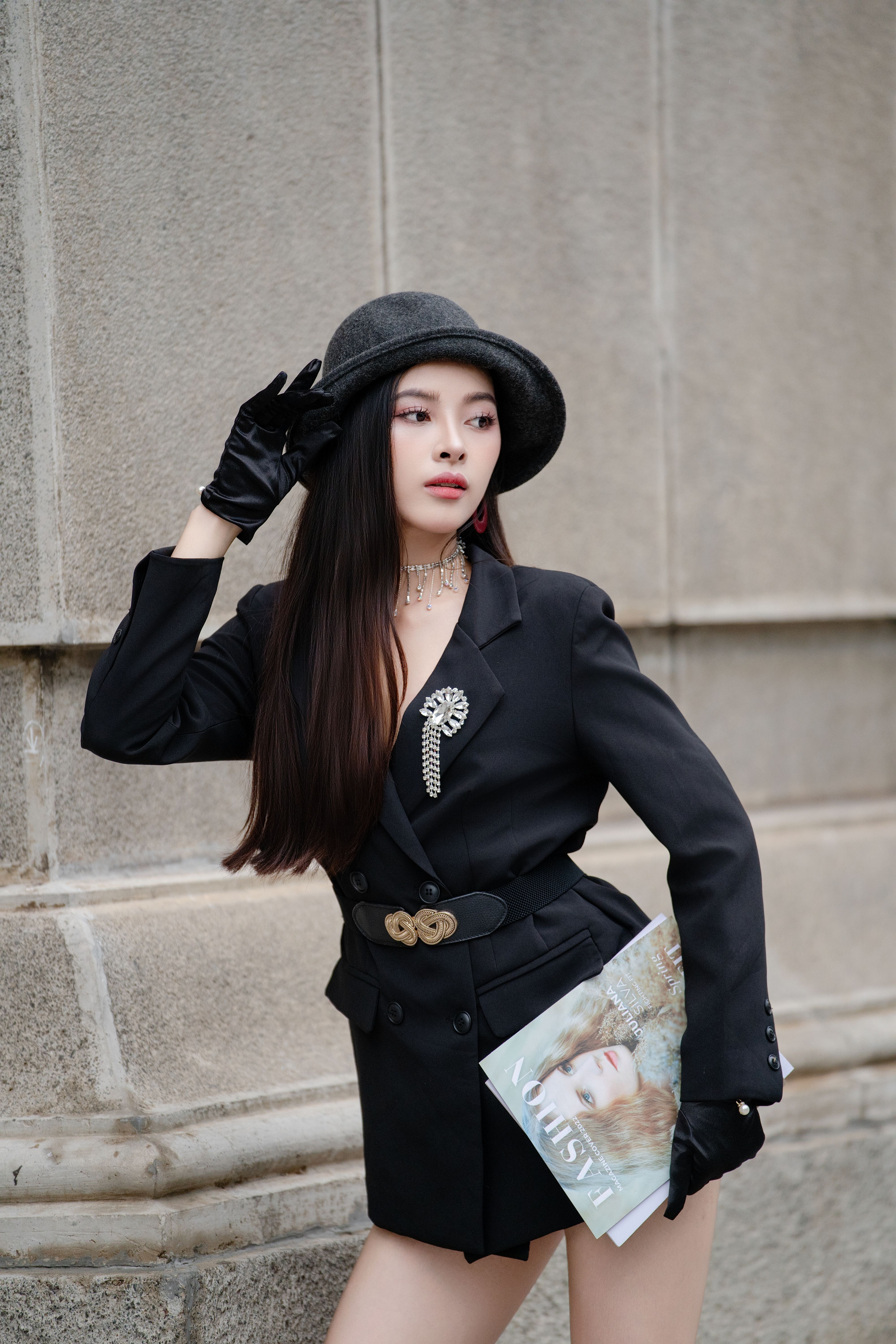 Elegant woman in black outfit with hat posing against urban backdrop.