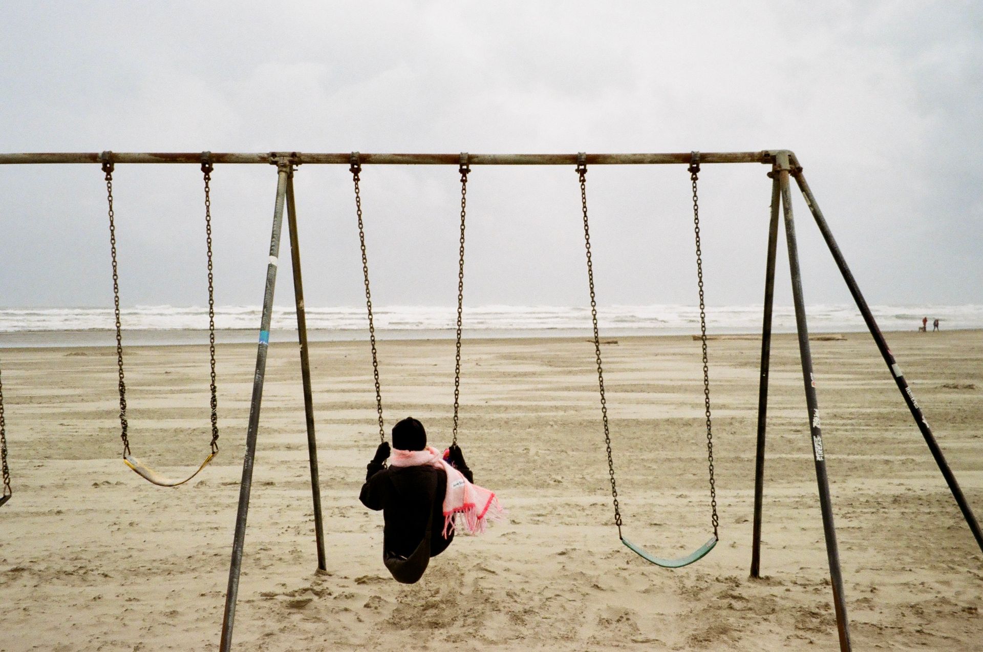 A person sits alone on a swing set by the ocean under an overcast sky.