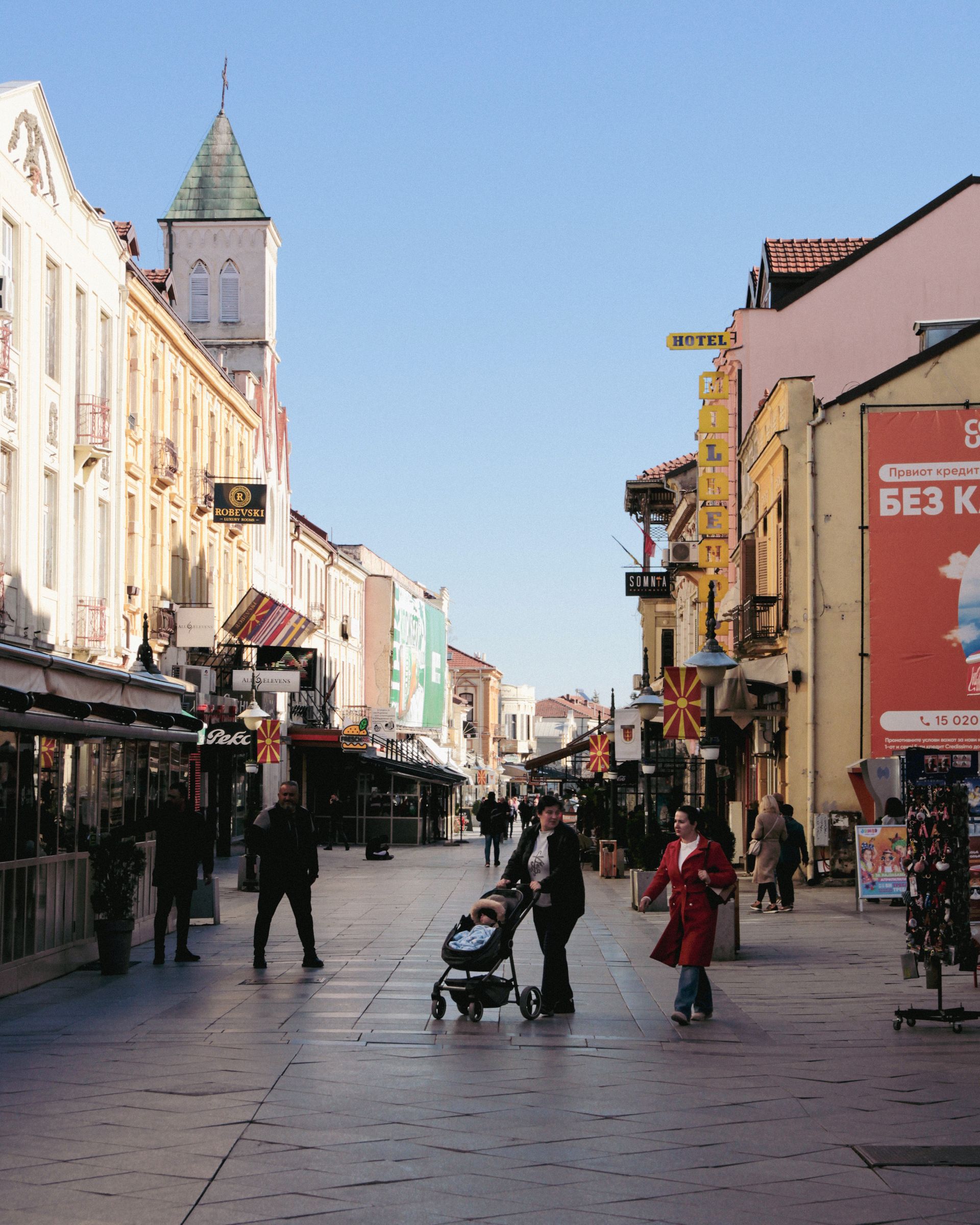 People strolling past café awnings and consular-era façades on Širok Sokak, with the Sacred Heart church tower and North Macedonian flags in Bitola