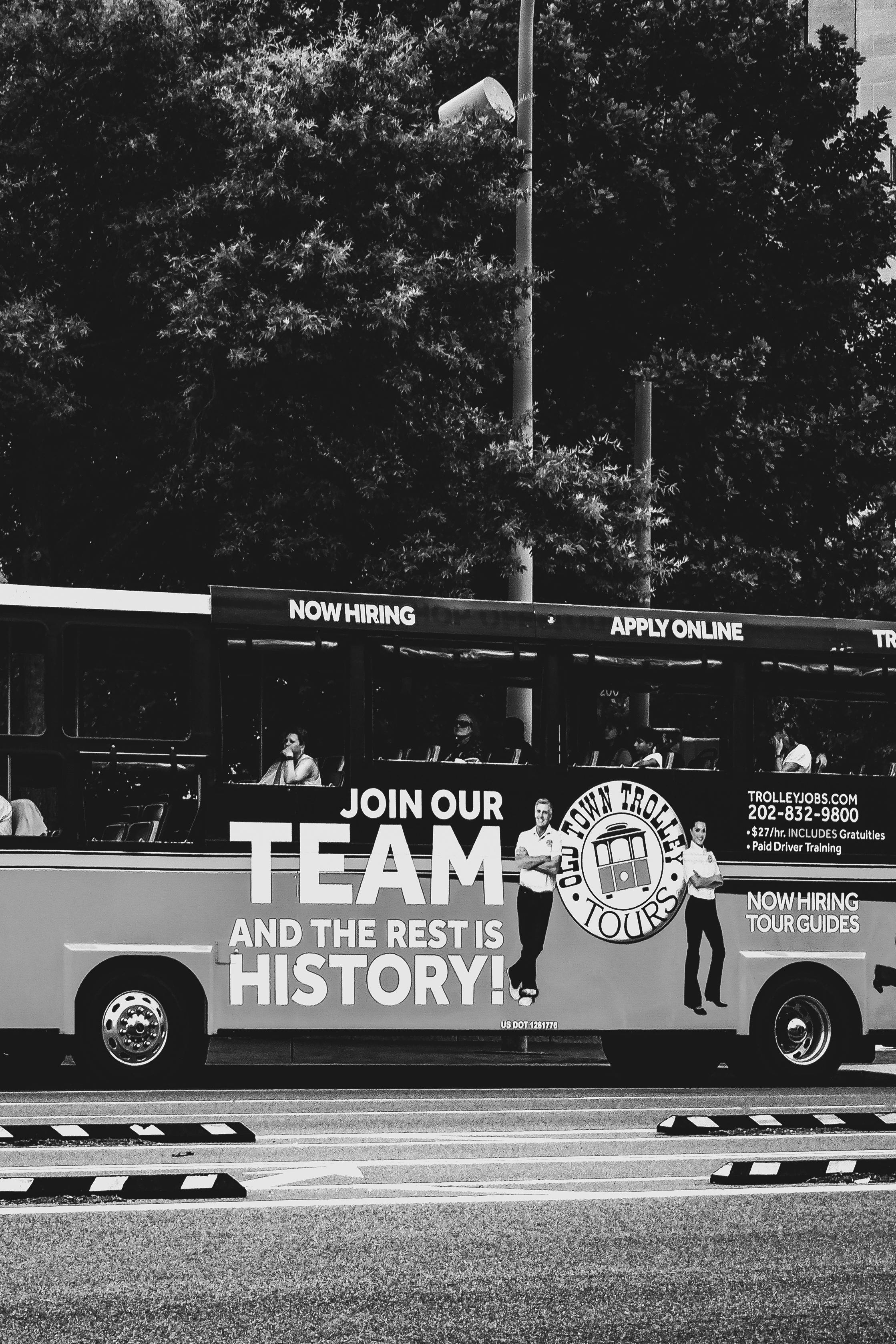 A black and white image of a tour bus with a recruitment advertisement in an urban setting.