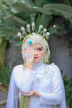 A bride in a white dress with intricate traditional headdress posing in a lush outdoor setting.