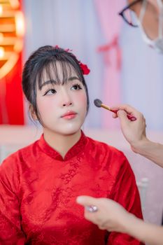 Asian woman in red dress having makeup applied, preparing for an event.