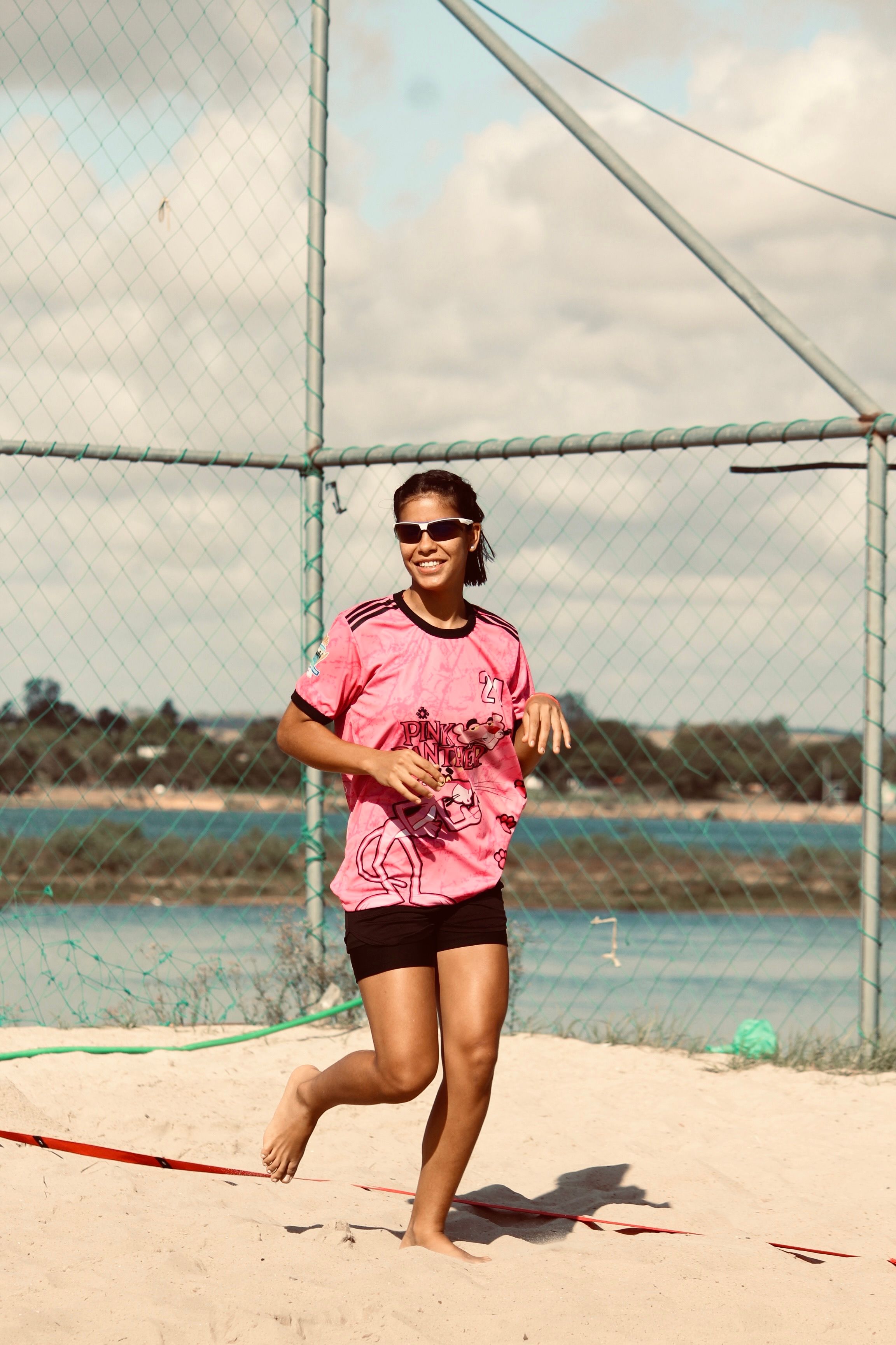 Smiling woman playing beach volleyball under sunny skies near a scenic water view.
