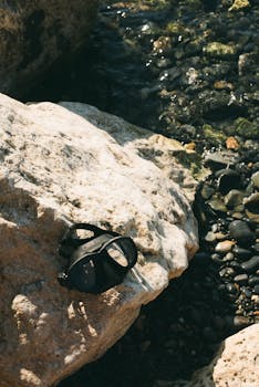 A diving mask resting on a sunlit rock beside clear coastal waters in Greece.