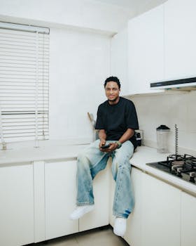 Casual man in black shirt sitting in a modern white kitchen holding a phone.