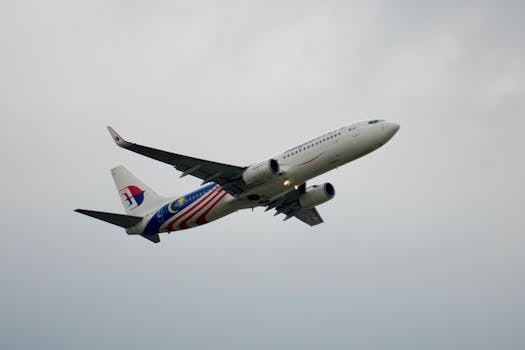 A Boeing 737 airplane adorned with the Malaysian flag livery flying against a cloudy sky.