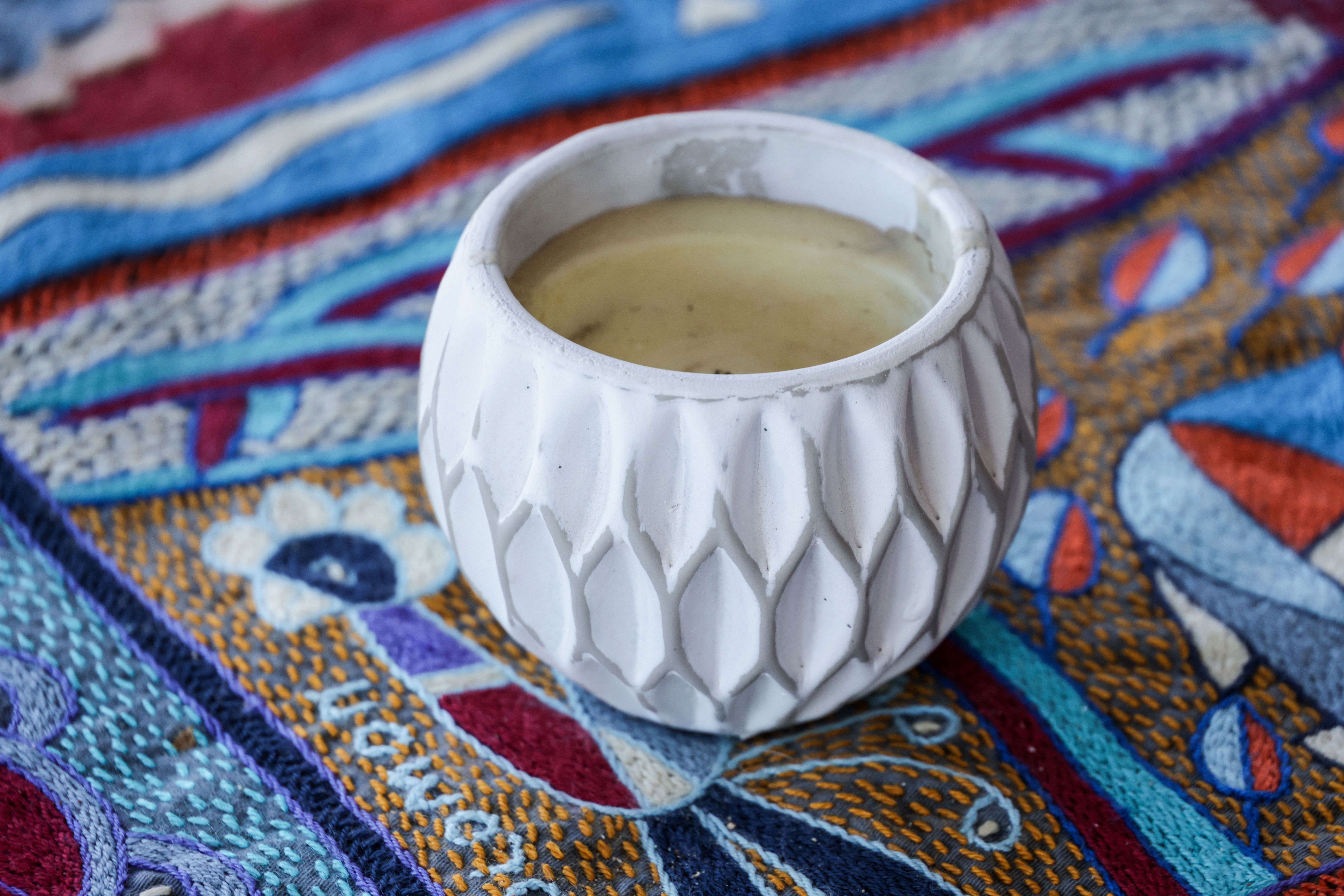 A white ceramic candle holder rests on a vibrant, textured fabric with intricate designs, adding aesthetic charm.