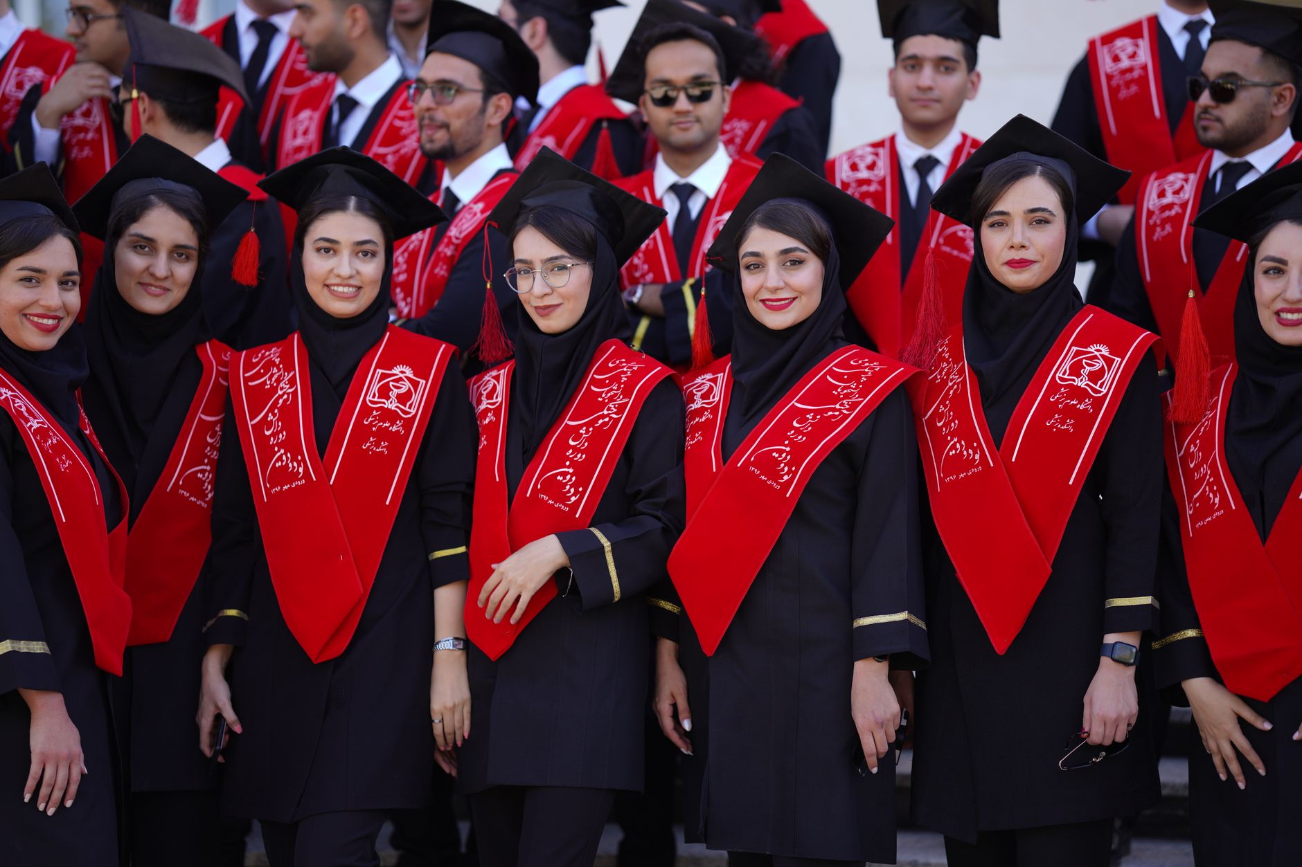 A group of Iranian students celebrating their graduation in traditional academic attire with red sashes.