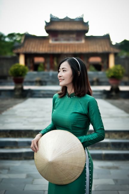 Serene woman in green Ao Dai with conical hat in front of Vietnamese temple.