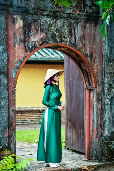 Woman in green Ao Dai and conical hat standing in historical archway, Vietnam.