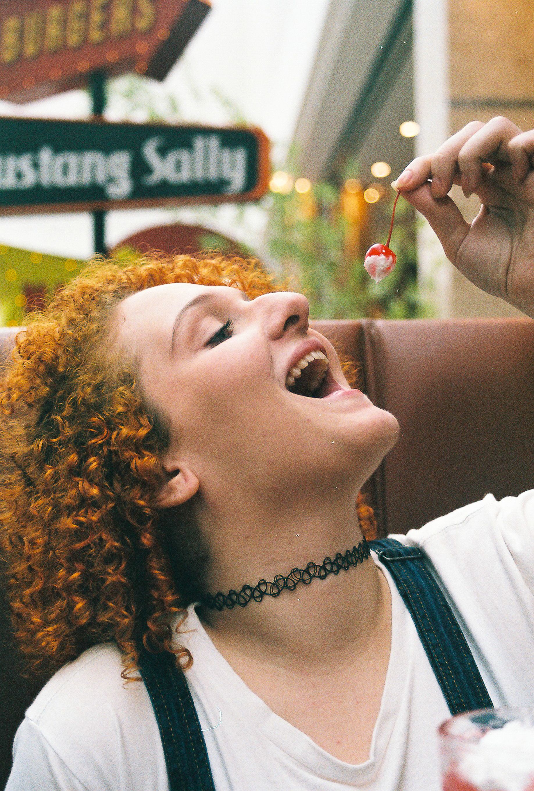 Smiling teen girl with curly hair savoring a cherry indoors at a retro-style diner.