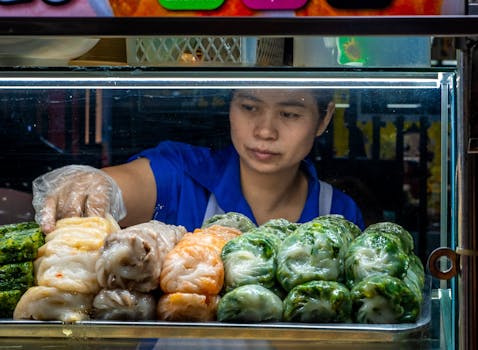 A street vendor arranges colorful steamed dumplings behind a glass food display in a market.