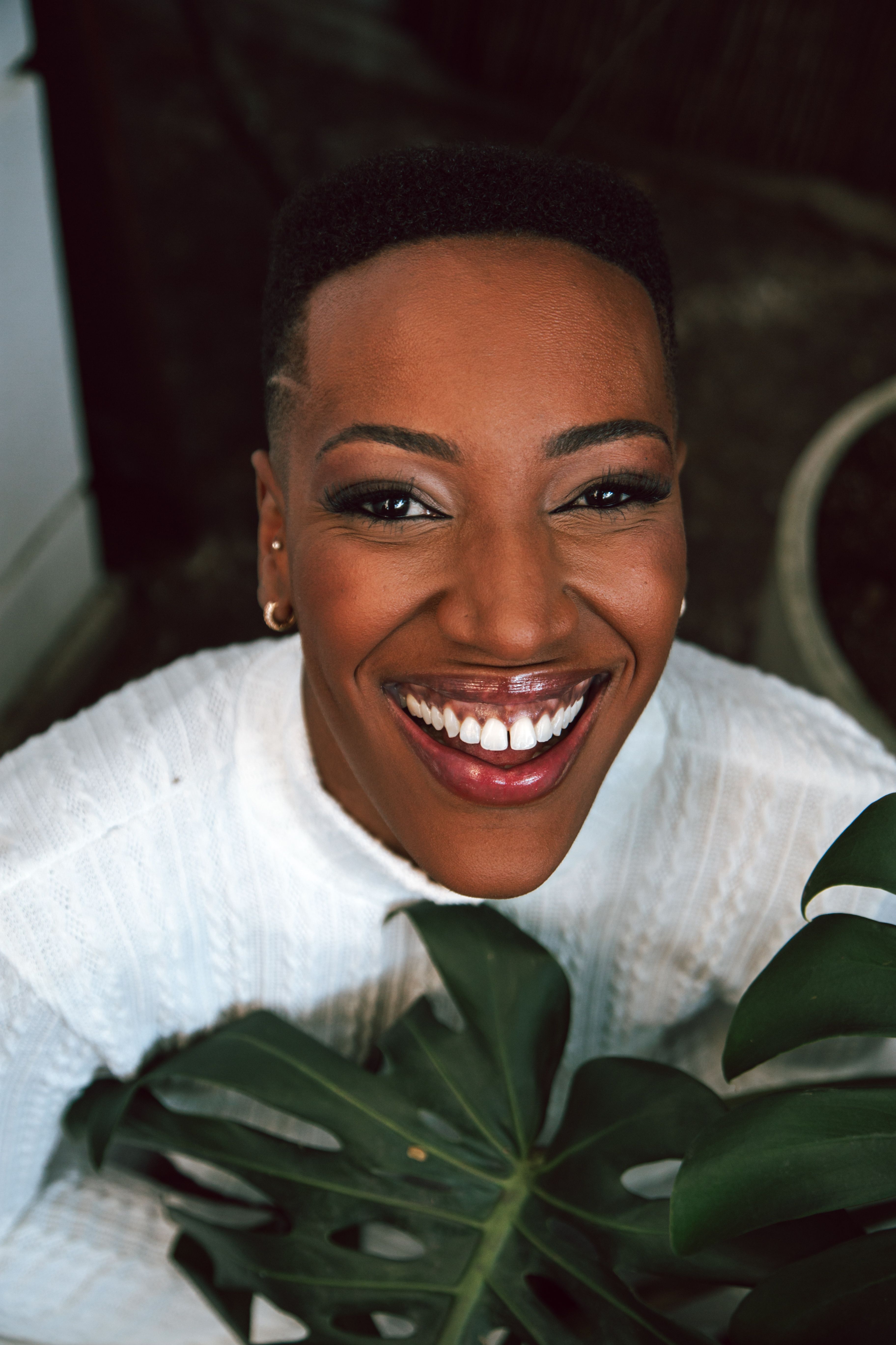 Cheerful woman in white top holding monstera leaf, smiling warmly indoors.