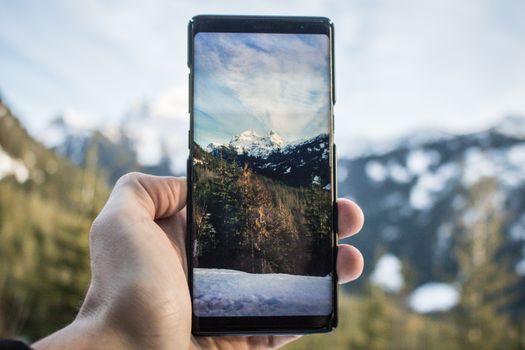 Hand holding a smartphone displaying a mountain view. Snowy peaks and forest seen through the screen.