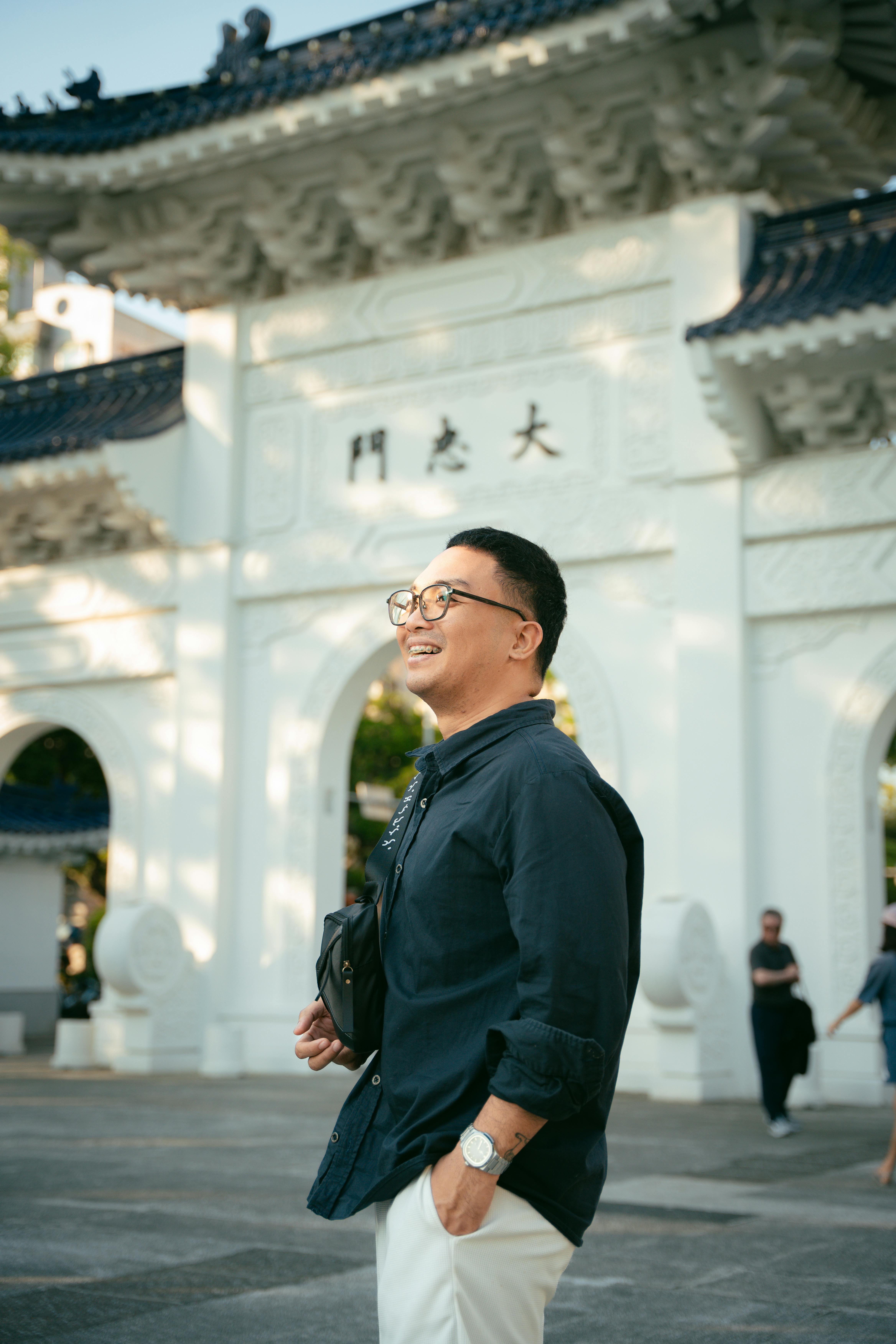 A man smiles in front of a traditional Asian architectural entrance, enjoying a sunny day.