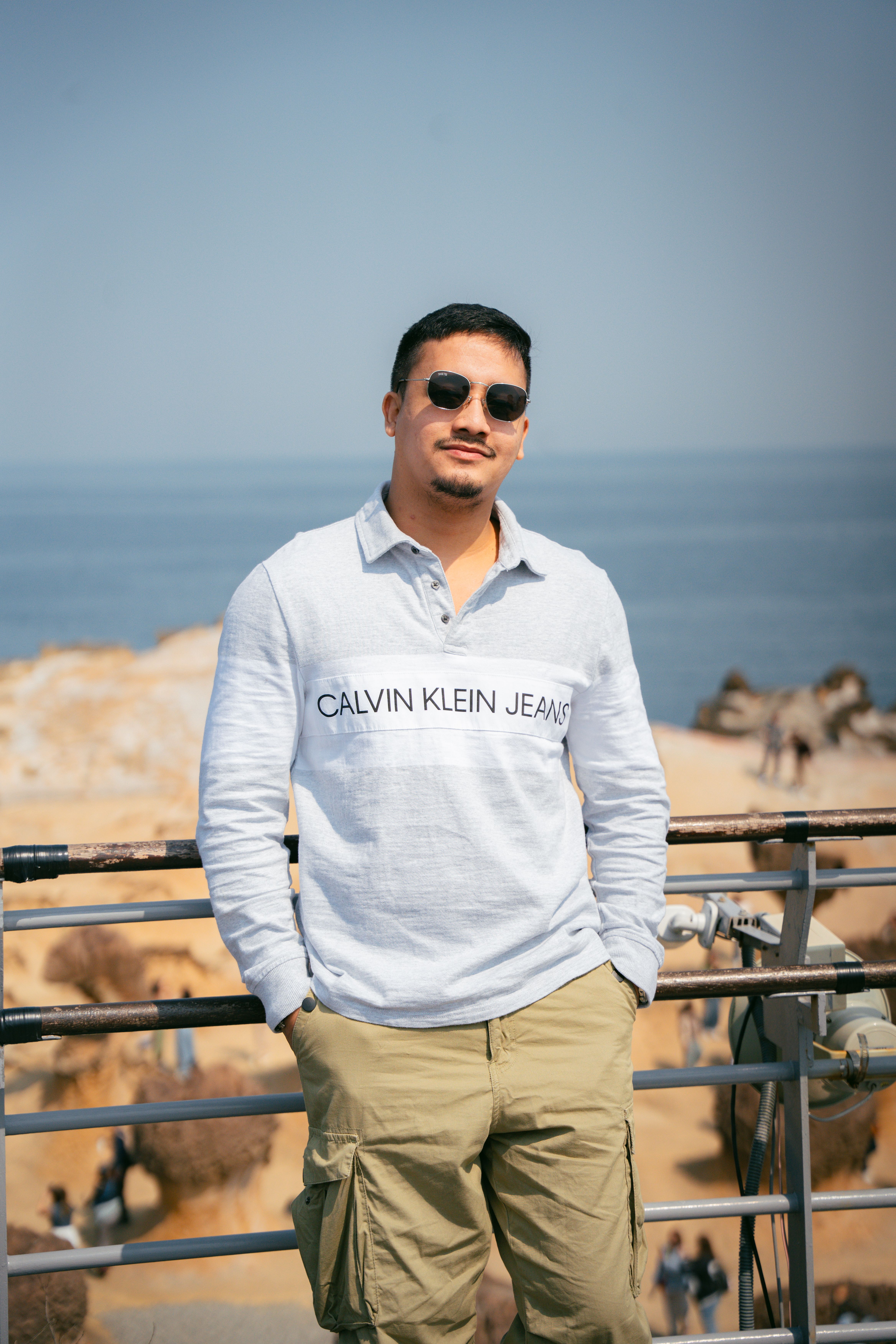 A man wearing sunglasses and casual clothes poses by the ocean on a sunny day.