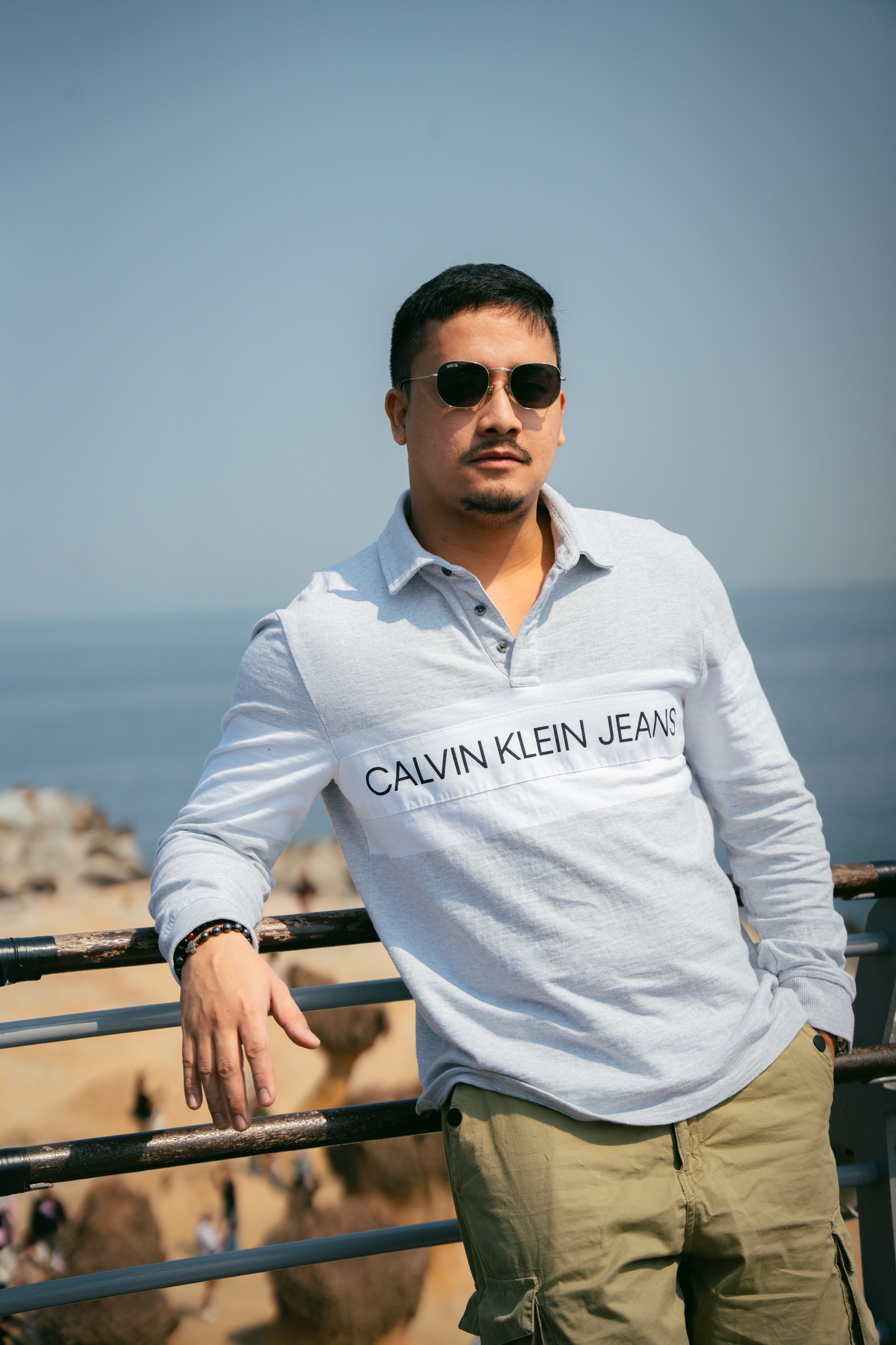 Casual man in sunglasses and branded clothing poses by the ocean.