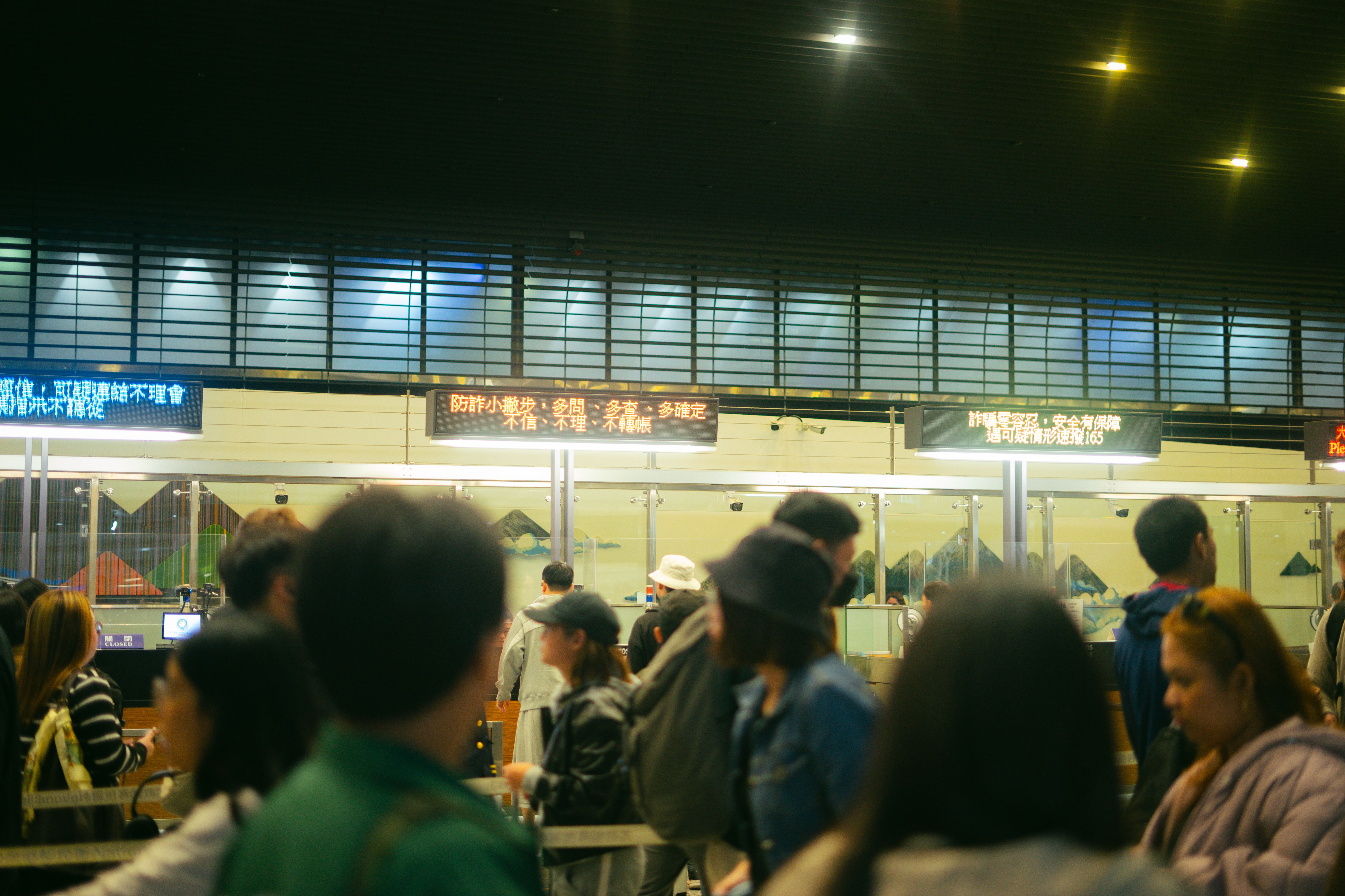Travellers queue at an immigration checkpoint beneath electronic information boards in an airport terminal