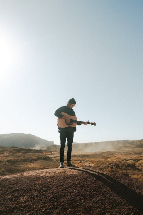 Free Full body male in warm clothes standing in endless field in morning sunlight and playing guitar under blue sky Stock Photo