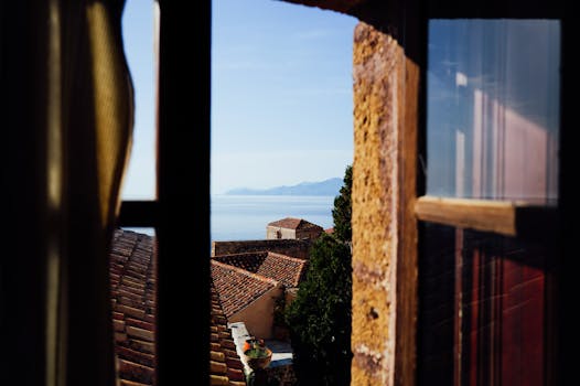 Charming Mediterranean coastal view through a rustic window with terracotta rooftops and sea horizon.