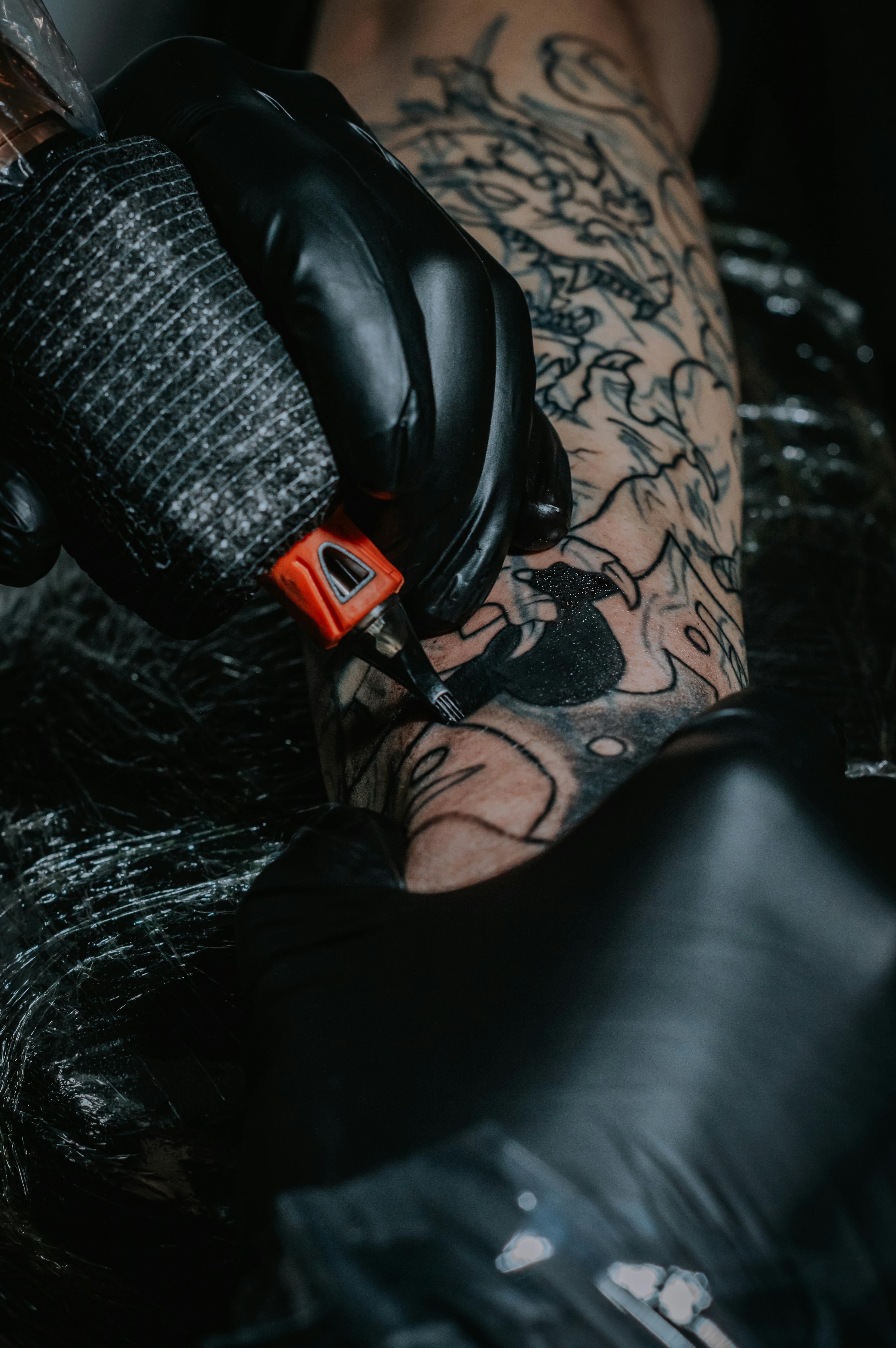 Intricate black tattoo being inked by a gloved artist, showcasing detailed artistry on arm.