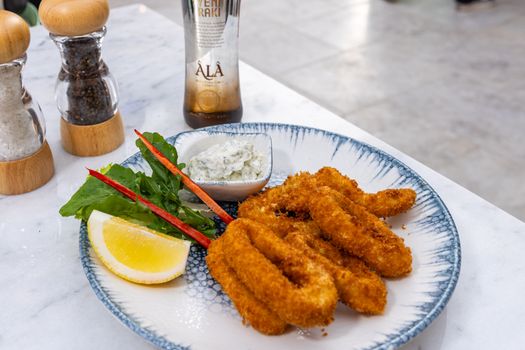 Golden calamari rings served with a wedge of lemon and dipping sauce on a marble table.