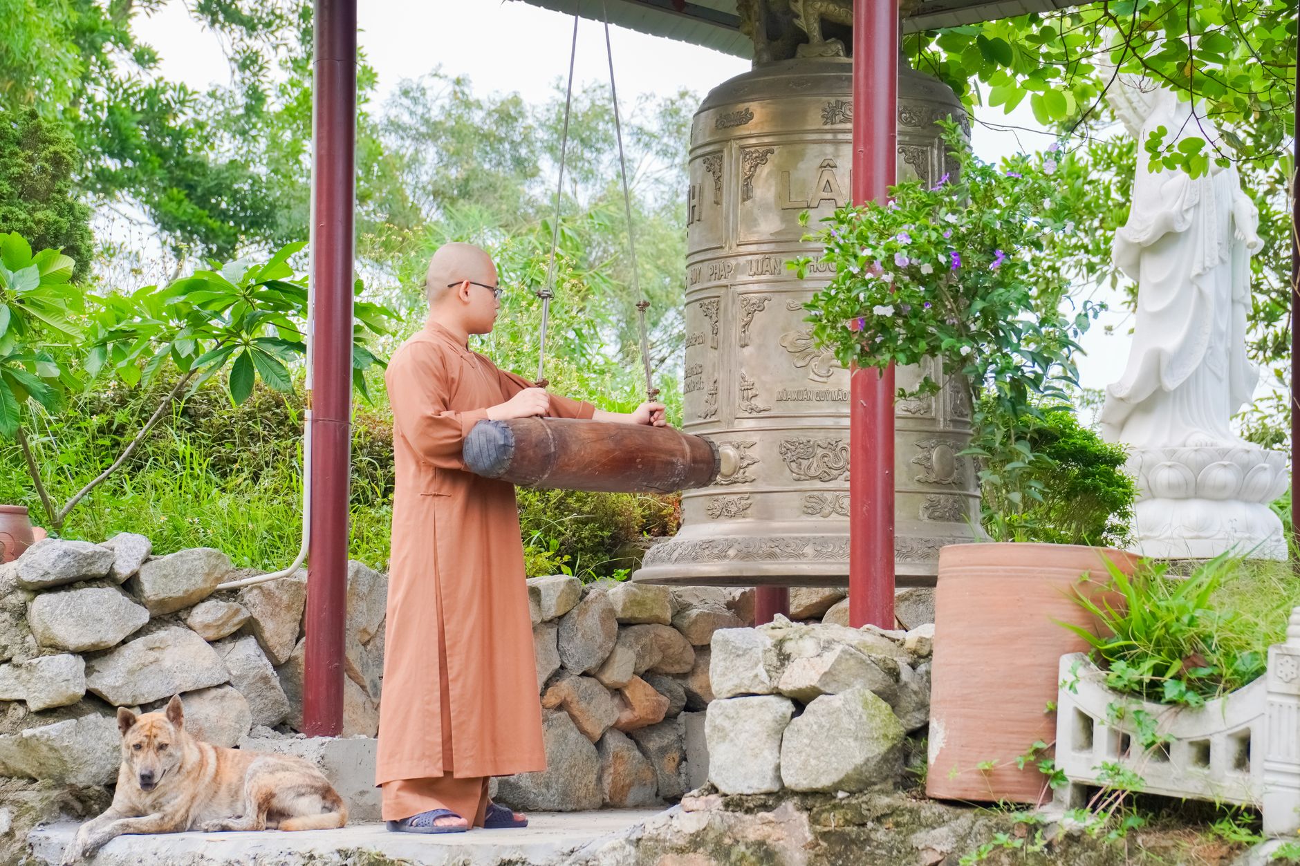 A Buddhist monk stands next to a large bronze bell in a temple garden in Thạch Lâm, Việt Nam.