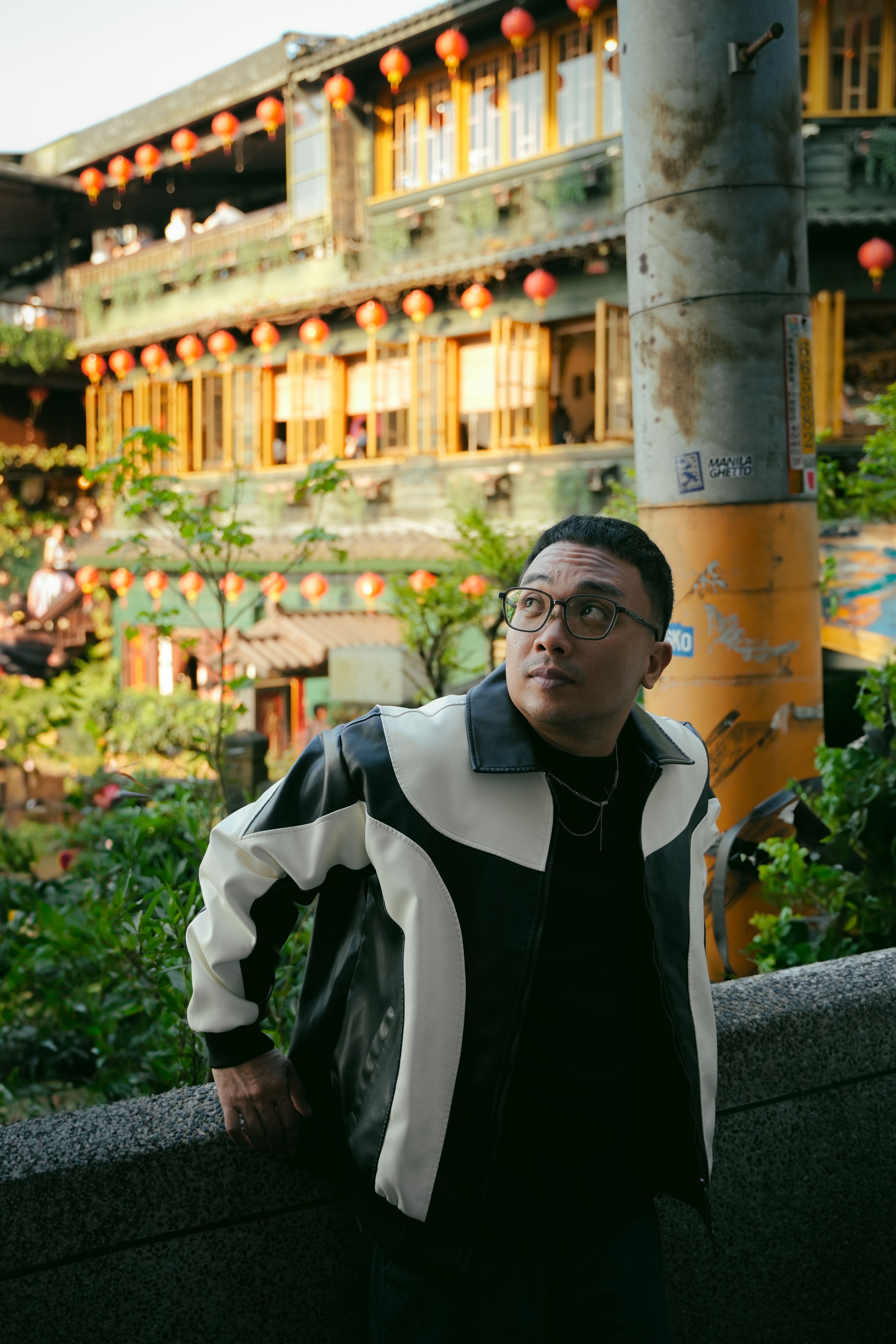 A man wearing a fashionable jacket gazes upwards in an Asian street setting with lanterns.