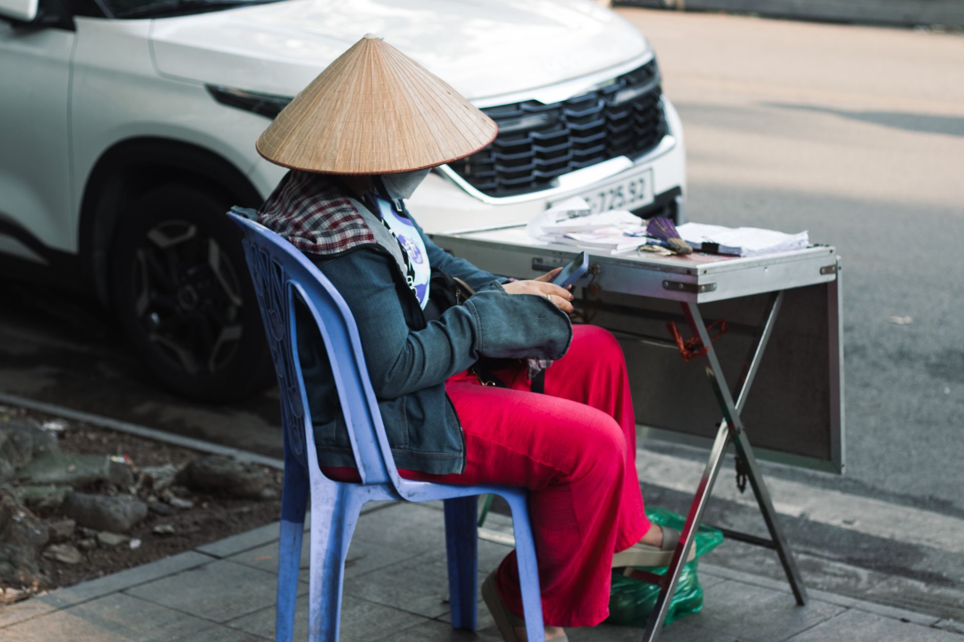 Street vendor with non la hat in Hanoi, Vietnam, using a phone by a road.
