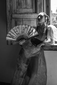 Artistic black and white portrait of a woman with a fan, evoking a moody and vintage atmosphere.