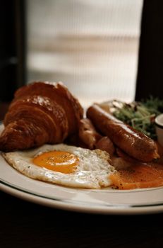 A hearty breakfast plate with croissant, sunny-side-up eggs, sausage, and salad.
