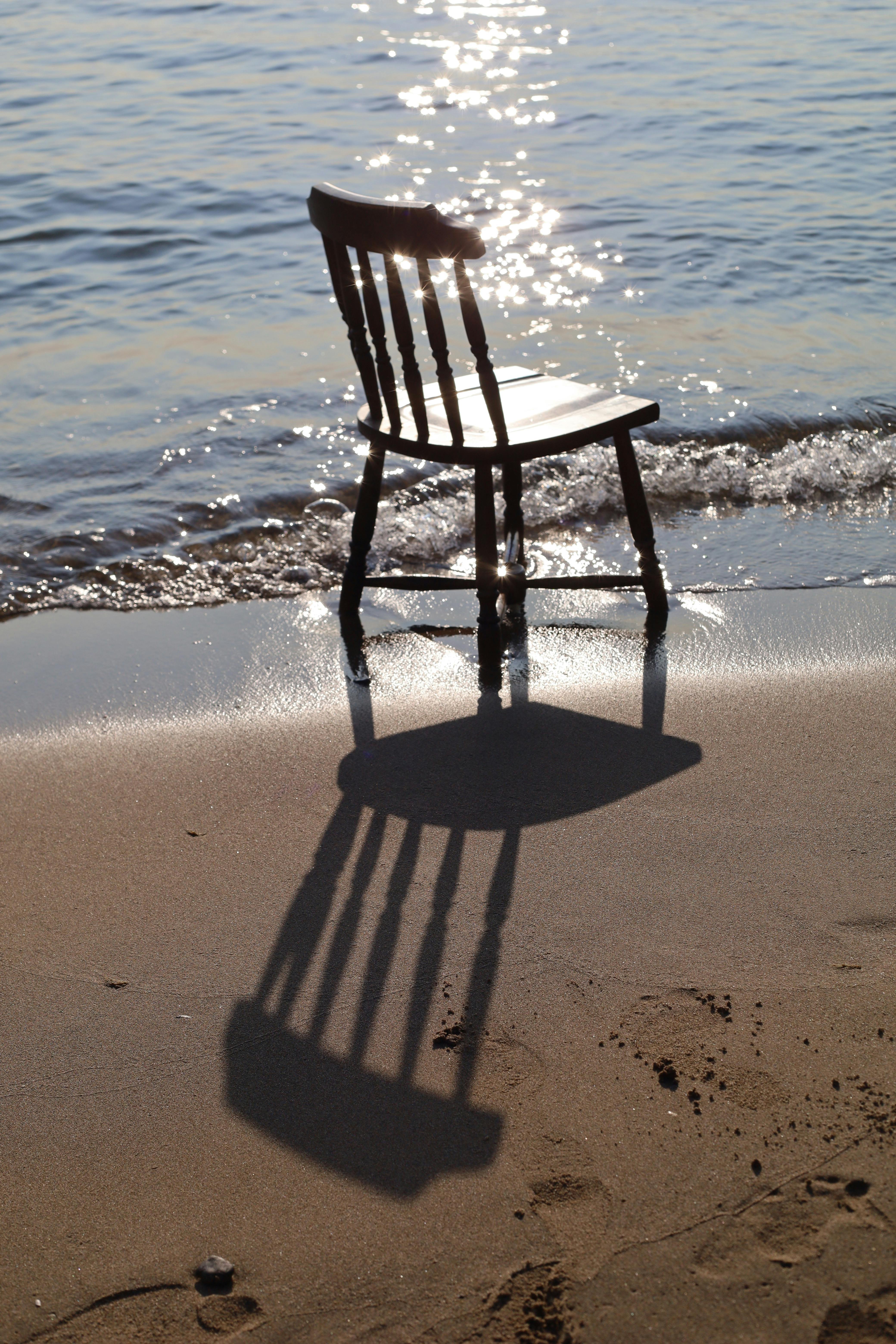 A solitary wooden chair casting a shadow on a sunlit beach by the water.