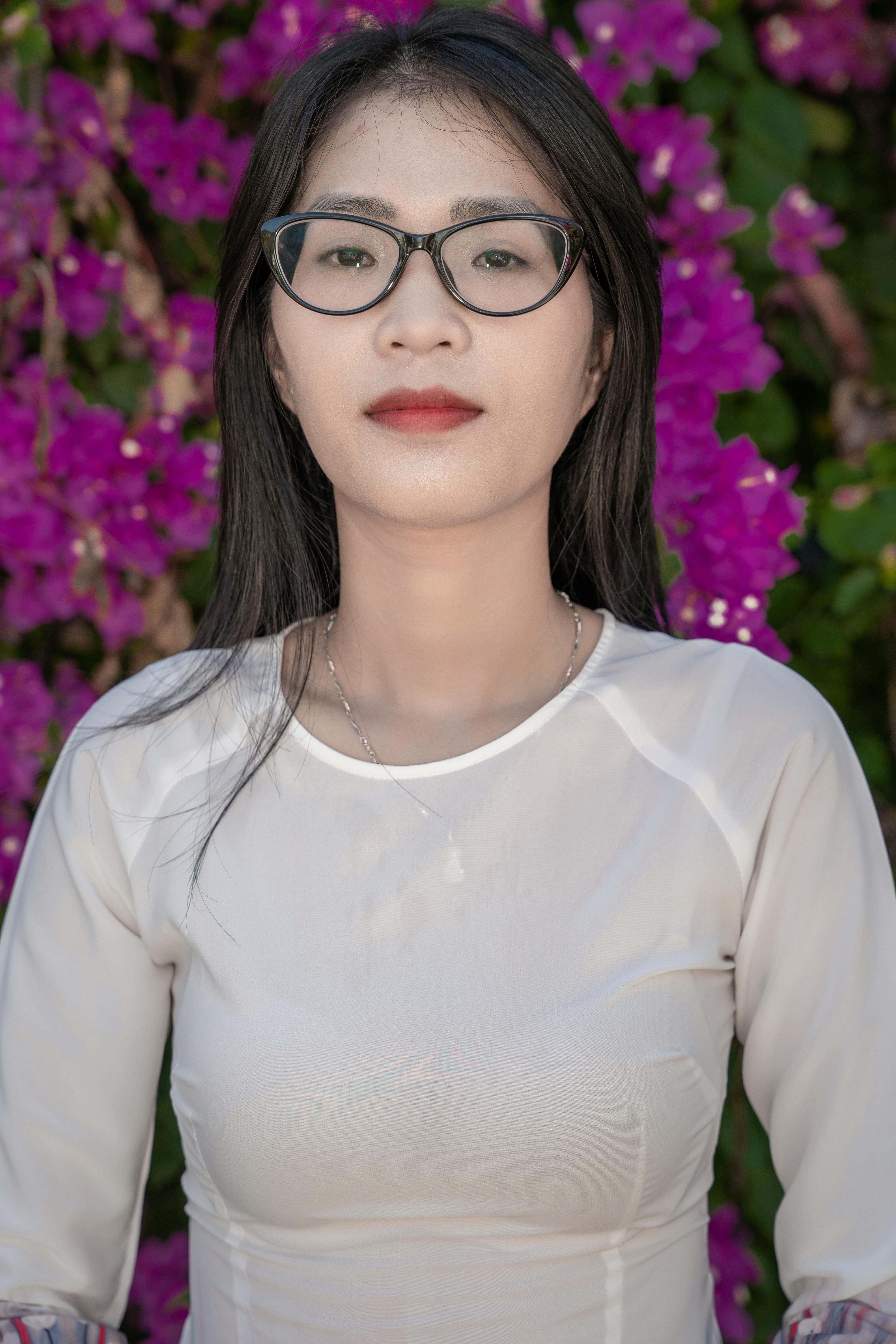 A woman with glasses poses confidently in front of vibrant purple flowers, showcasing elegance and natural beauty.
