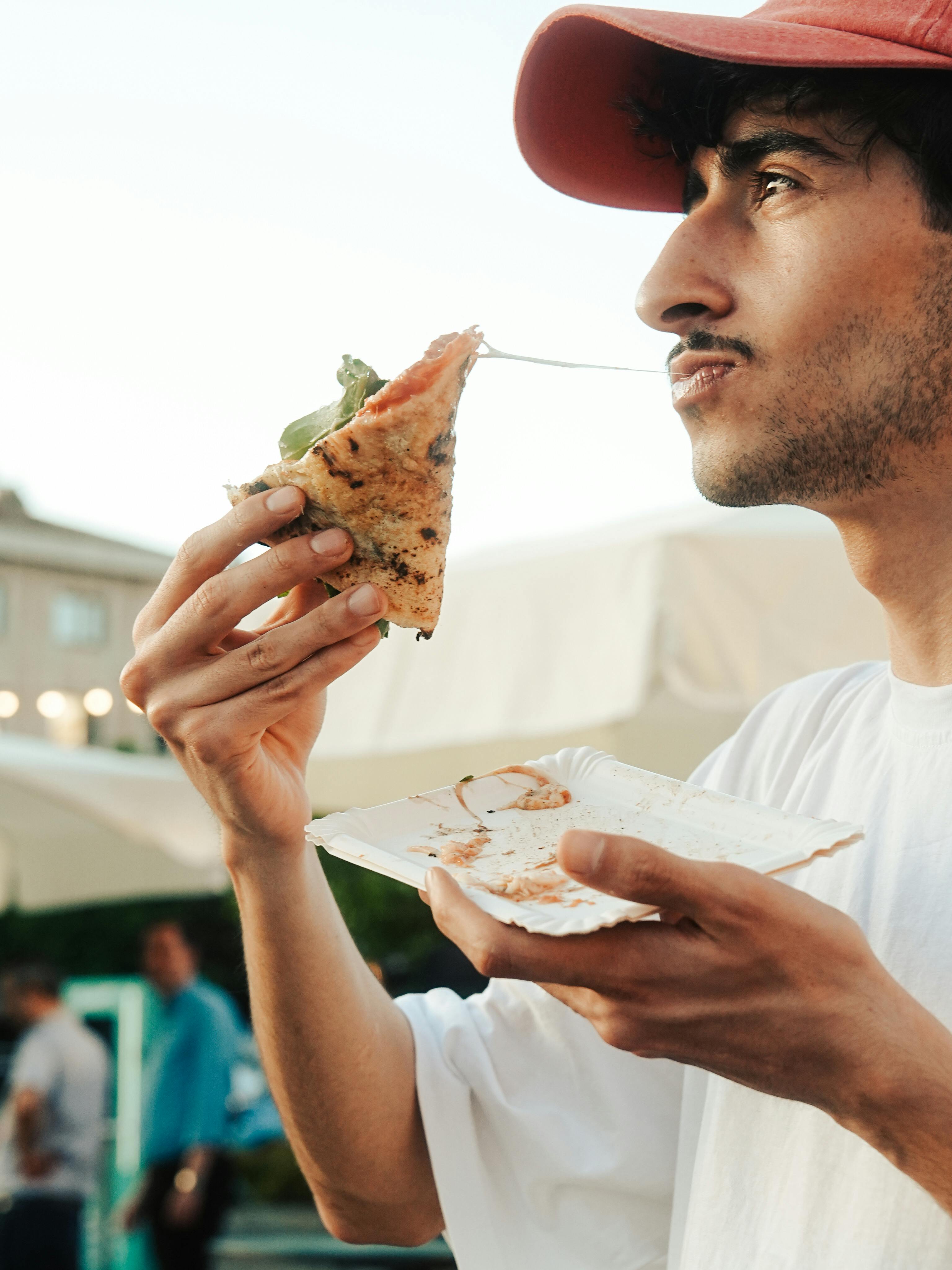 Free Young man savoring a delicious slice of pizza in an outdoor setting, conveying enjoyment and leisure. Stock Photo