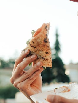 Close-up of a hand holding a pizza slice outdoors during summer.