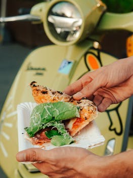 Hand holding a fresh pizza slice near a Vespa scooter, outdoors.