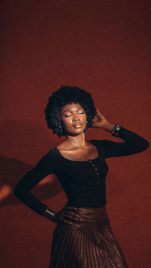 Free Stylish portrait of a woman with afro hair in a brown skirt, eyes closed against a rich red backdrop. Stock Photo