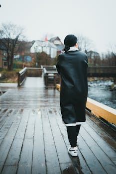 Back view of an adult wearing a black coat walking on a wooden bridge on a rainy day.