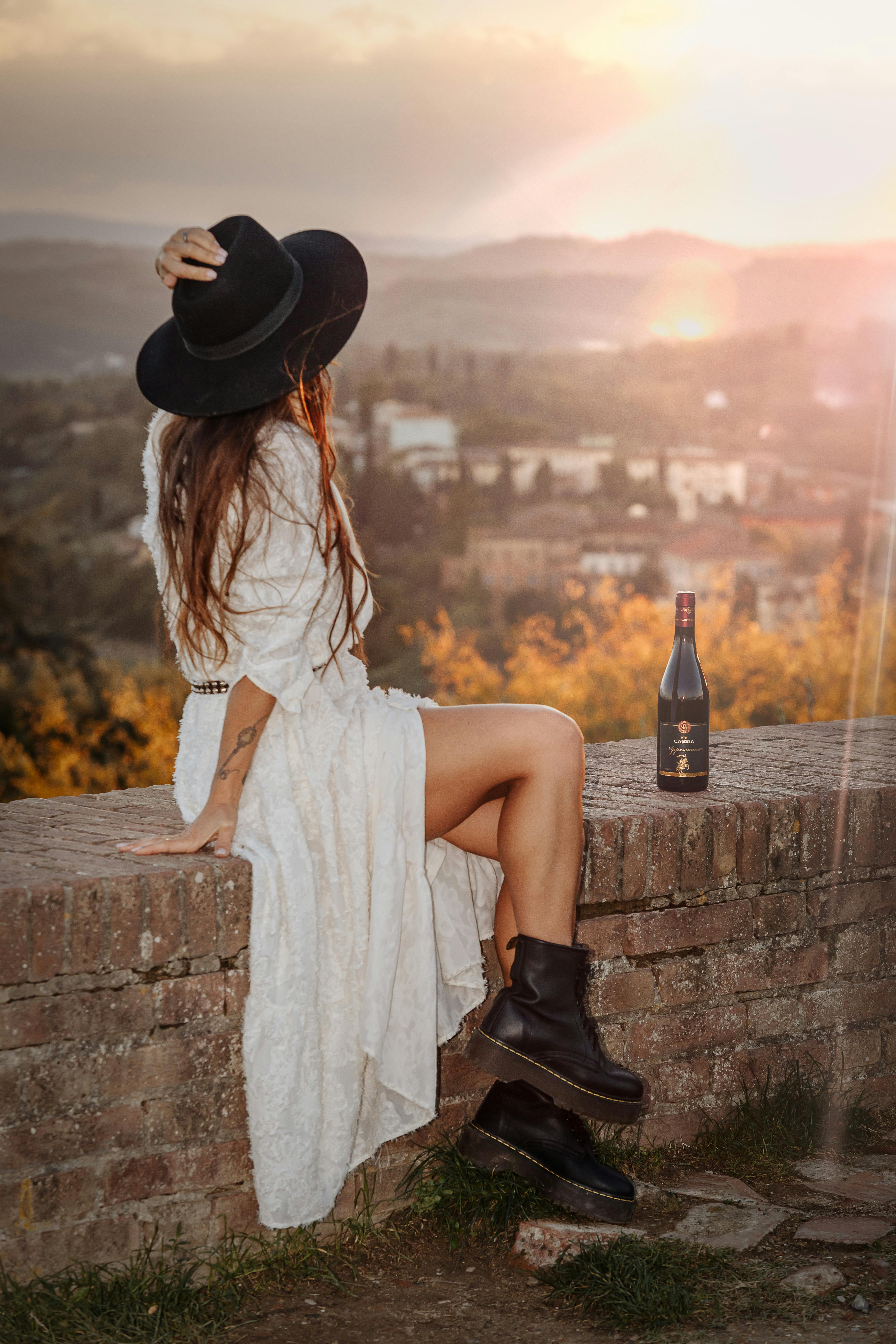 Woman in white dress and black boots relaxes with wine at sunset on brick wall, scenic view.