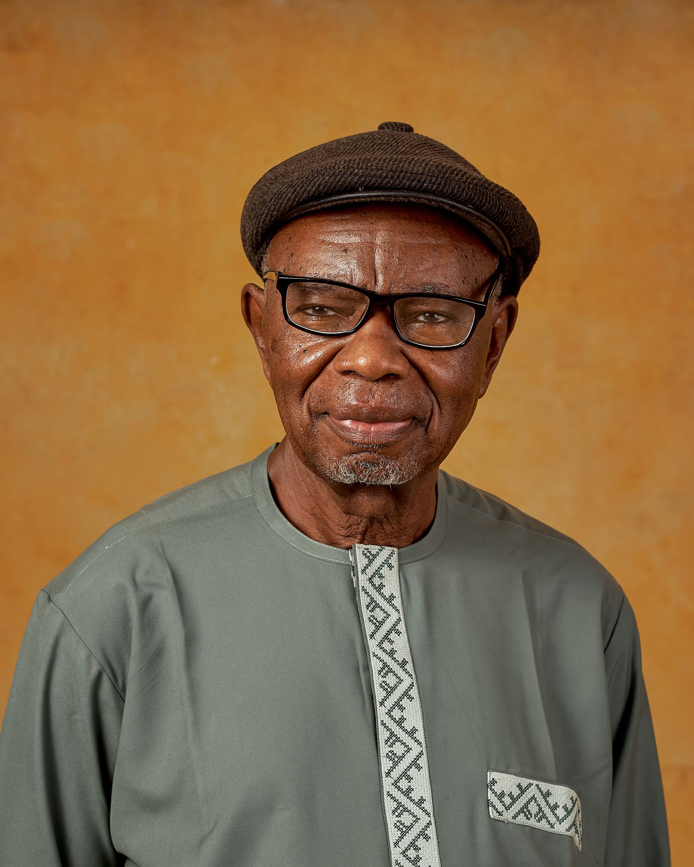 Elderly man wearing traditional outfit and cap, posing against a warm background.
