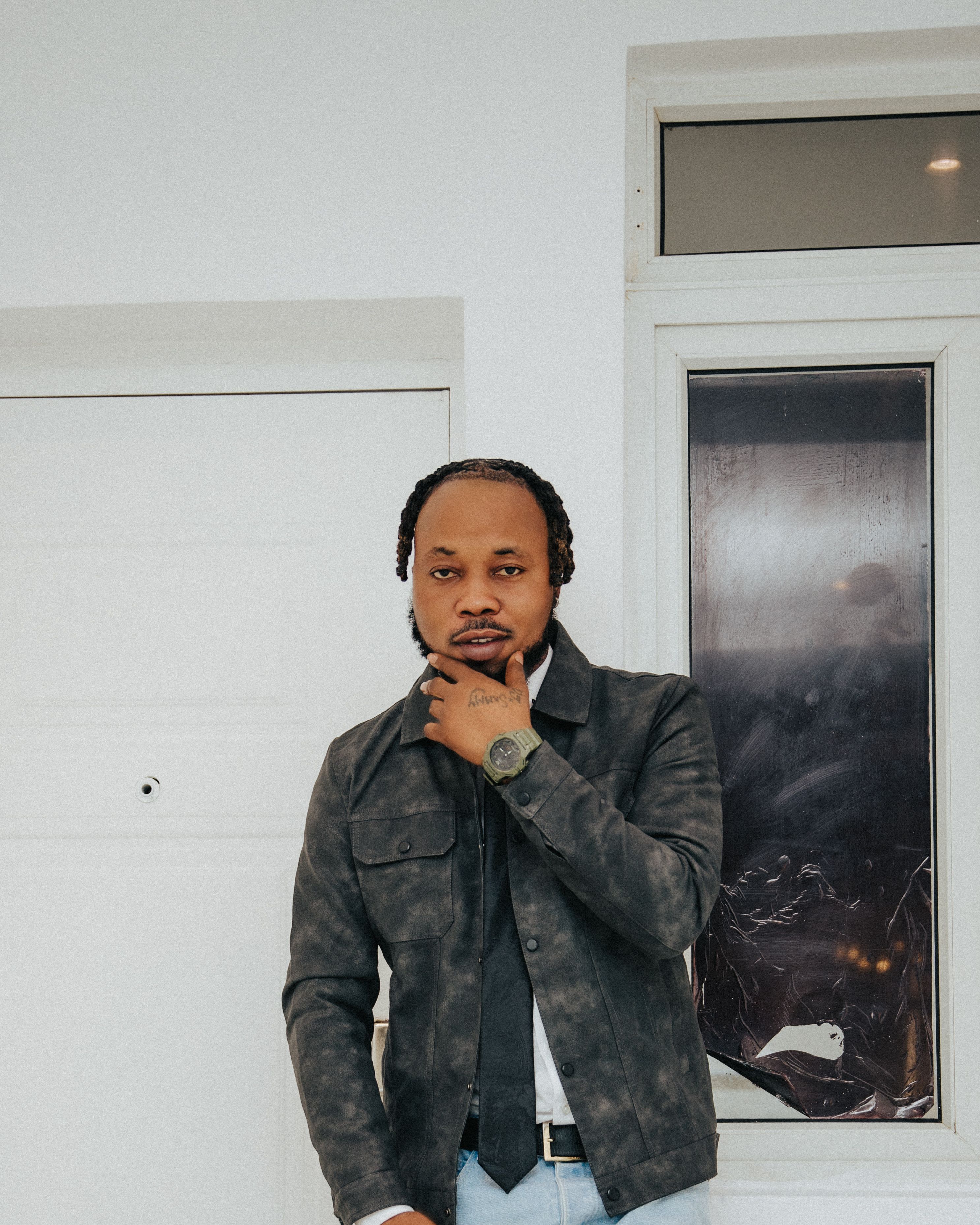 Man in black jacket and watch posing near white wall indoors with a thoughtful expression.
